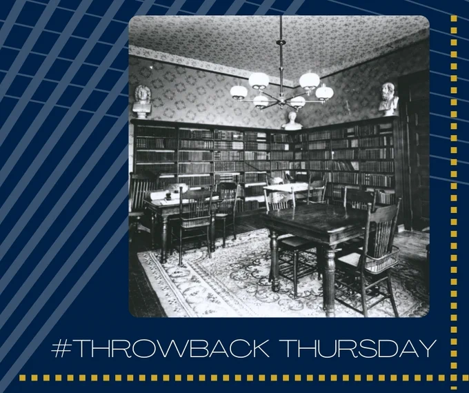 A black and white photograph of a small library with dark books on dark shelves and dark tables and chairs on a light patterned rug.