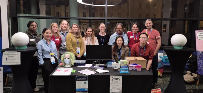 Photograph of the 'Would you get sick for science?' team stood behind their stand at the Imperial Lates event. There are various science engagement activities and objects on the tables.