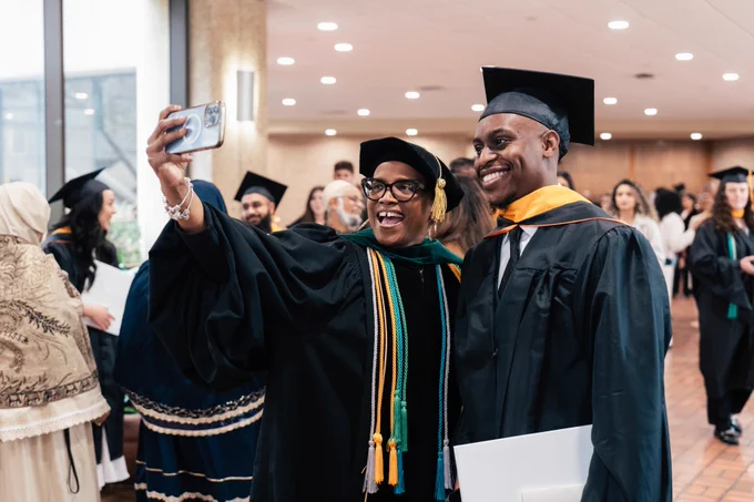 Two graduates in cap and gown taking a selfie with a smartphone during their graduation ceremony.