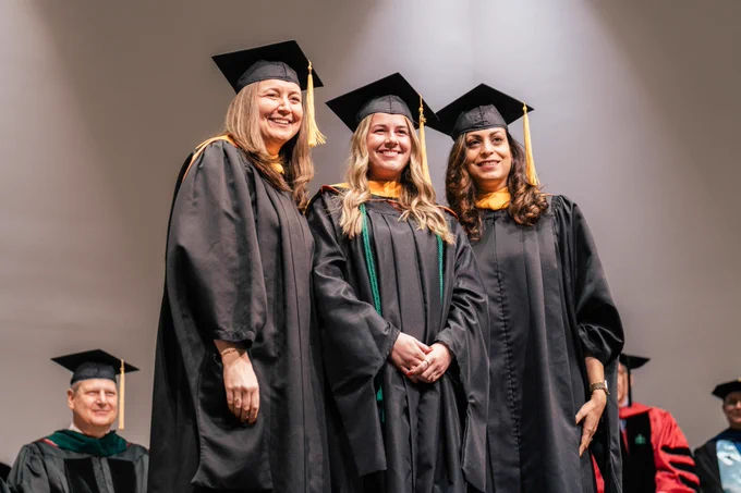 Three graduates wearing caps and gowns smiling proudly at a graduation ceremony.
