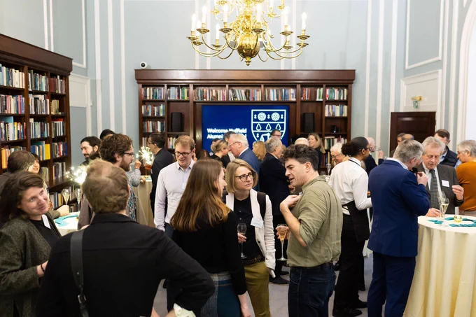A wide shot of a blue Georgian room with many small groups of adults of varying ages conversing. In the background is a screen saying 'Welcome Nuffield alumni' and dark bookcases line the walls.