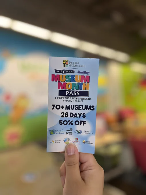 Closeup of a hand holding a Museum Month pass with a blurred reading room and Seuss mural in the background at the Denny Sanford Children’s Library at San Diego Central Library.