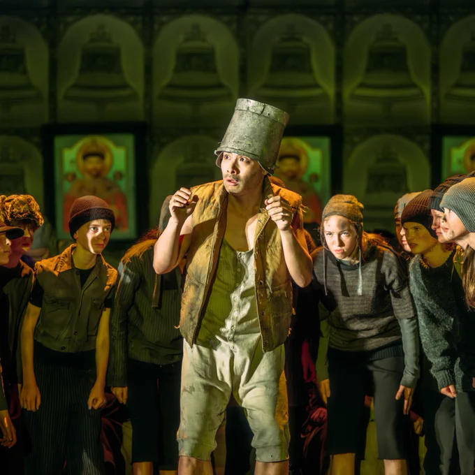 A man playing in Boris Gudunov with a silver bucket on his head. He looks scared and has a circle of children around him.