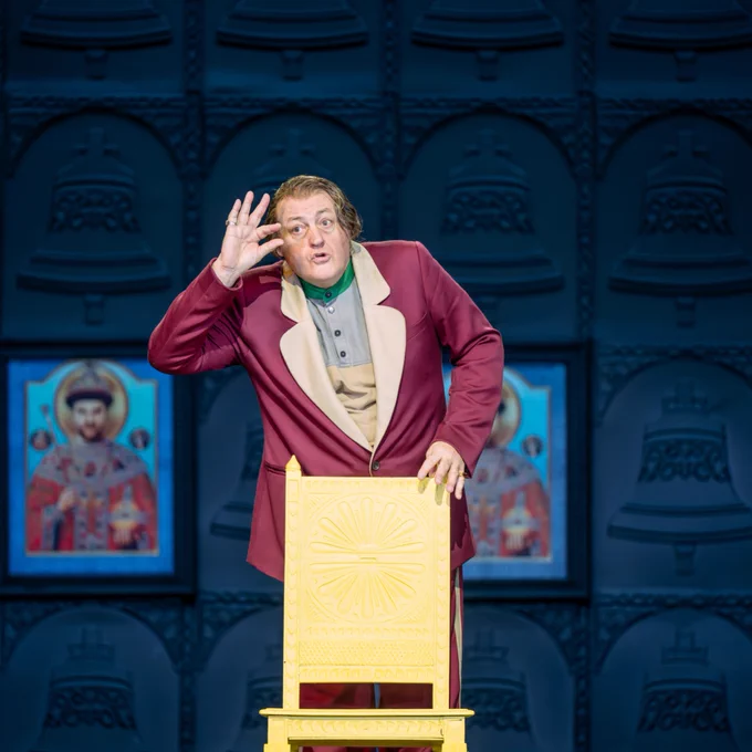 A man with a magenta jacket on with an expressive face, stood behind a yellow chair.