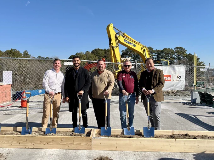Five people participating in a groundbreaking ceremony with shovels, standing in front of a excavator at a construction site.