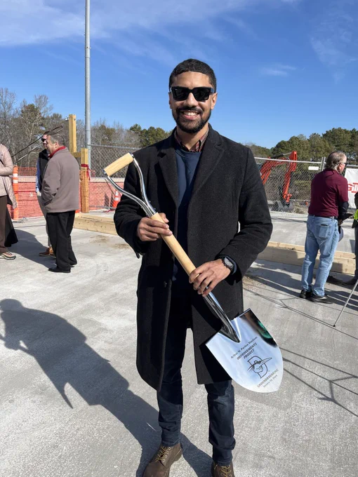 Person wearing sunglasses and a coat, smiling and holding a ceremonial shovel at a groundbreaking event.