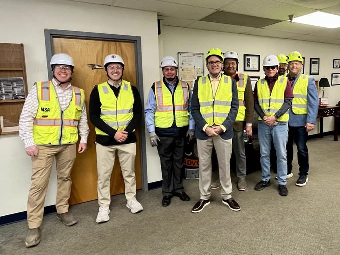 Group of six individuals in safety gear including helmets and high-visibility vests, standing indoors.