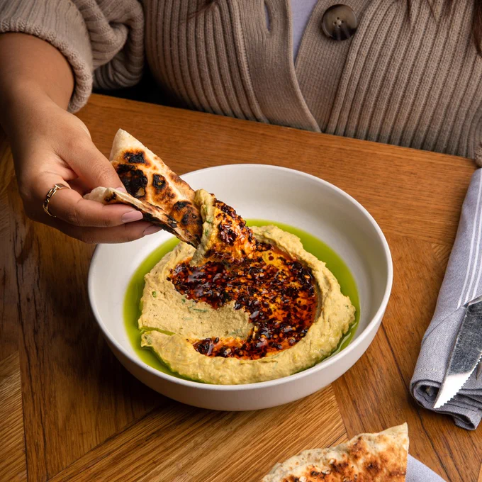 Hand dipping toasted pita bread into a bowl of creamy hummus drizzled with olive oil and spicy chili oil on a wooden table.