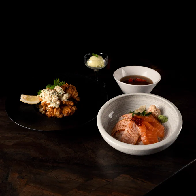 Bowl of fresh salmon sashimi with roe and wasabi, served alongside a small bowl of soy sauce and a plate of fried chicken topped with creamy sauce and a lemon wedge.