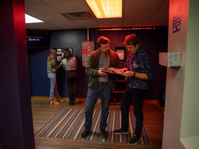 Two individuals examine a book together in a dimly lit escape room, with others in the background.