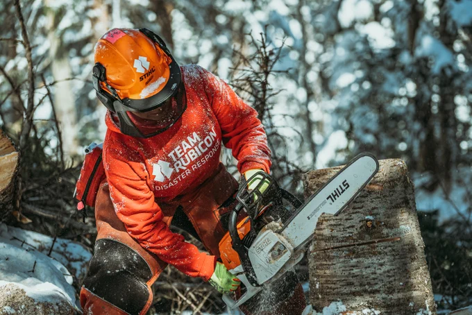 Person wearing orange safety gear and helmet using a chainsaw to cut a snow-covered log in a forest.