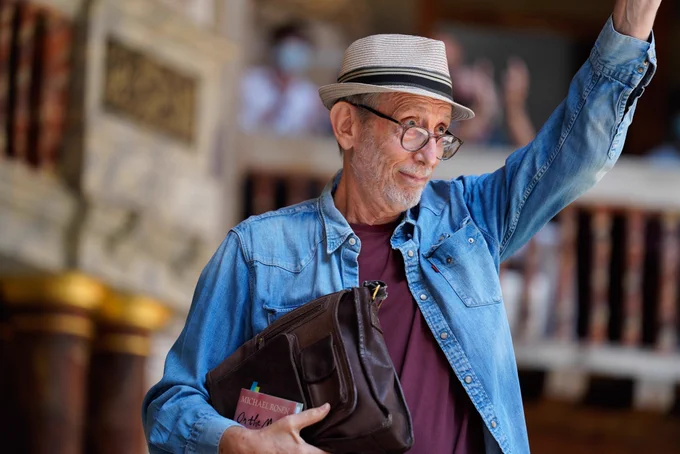 Photograph of Michael Rosen. Take at Shakespeare's Globe. He is wearing a hat, waving to the crowd and carries a copy of his book On the Move.