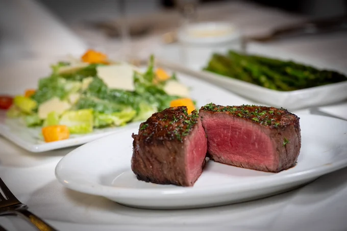 Medium-rare filet mignon steak sliced in half on a white plate, garnished with herbs, with a fresh Caesar salad and grilled asparagus in the background.