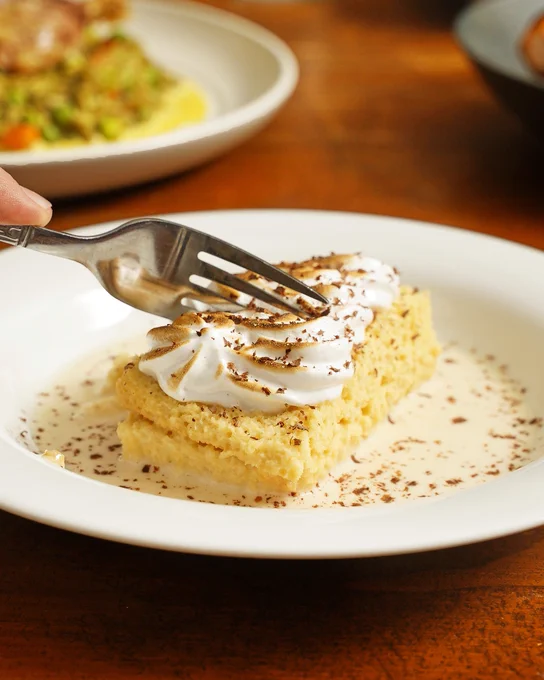 Fork cutting into a slice of tres leches cake topped with whipped cream and chocolate shavings on a white plate with creamy sauce.