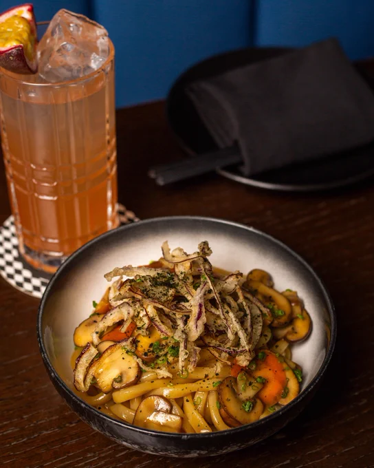 Bowl of udon noodles with sautéed mushrooms, carrots, and crispy fried onions on top, served on a dark wooden table.