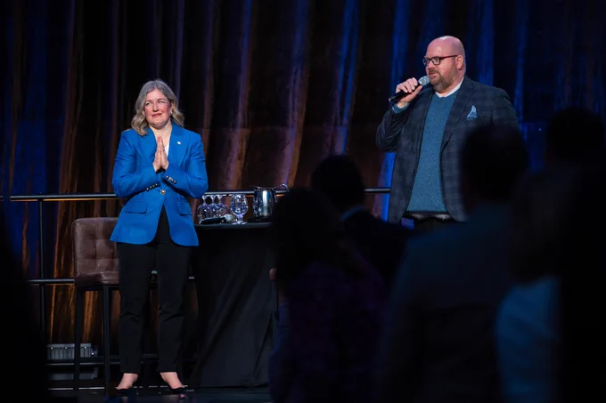 A person in a blue blazer stands on stage with hands clasped, next to another person speaking into a microphone. A table with glassware is behind them, and an audience is visible in the foreground.