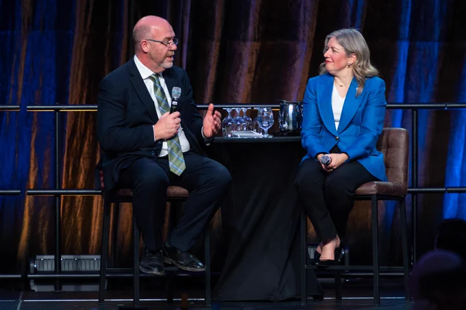Two people are seated on a stage engaged in conversation. One is holding a microphone and gesturing while the other listens attentively. They are dressed formally and seated at a small table holding water glasses. The backdrop features a dark curtain, creating a professional atmosphere.