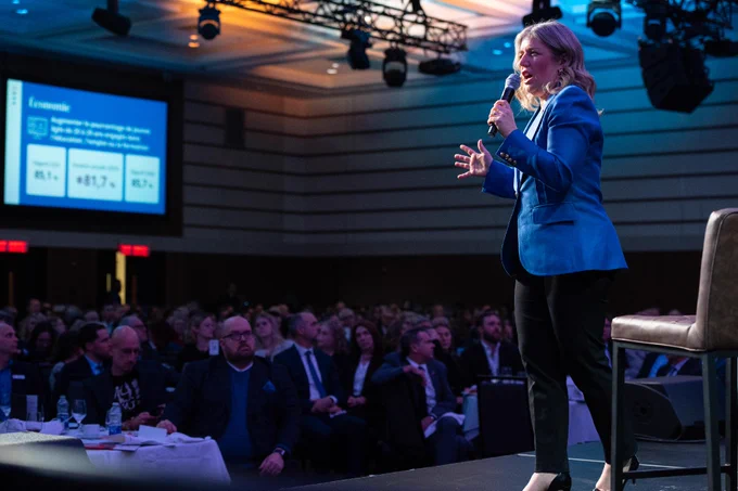 A person in a blue blazer speaks on stage with a microphone at a conference. The audience is seated, and a large screen in the background displays economic statistics. The room is well-lit, with a high ceiling and multiple lights and speakers overhead.