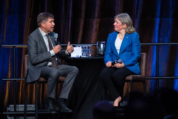 Two people are sitting on chairs at a round table, engaged in conversation. The person on the left is holding a microphone. Both individuals are dressed in formal attire, with the person on the right wearing a blue blazer. Behind them is a dark, draped backdrop.