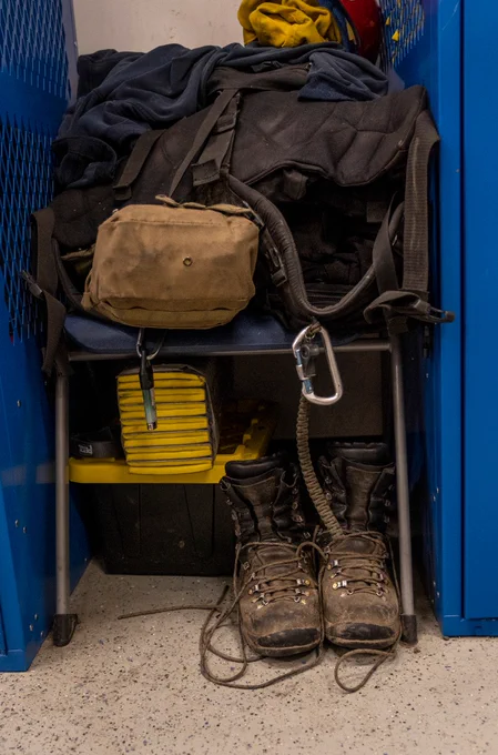 Worn hiking boots with untied laces placed under a chair holding a tan bag and black jacket inside a blue locker.