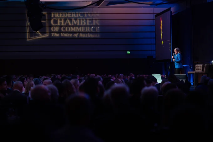 A woman speaks on stage to a large seated audience at a formal event. The Fredericton Chamber of Commerce logo is projected on the wall behind her. The room is dimly lit, except for the stage lighting.
--
Une femme s'adresse sur scène à un large public assis lors d'un événement officiel. Le logo de la Chambre de commerce de Fredericton est projeté sur le mur derrière elle. La salle est faiblement éclairée, à l'exception de l'éclairage de la scène.