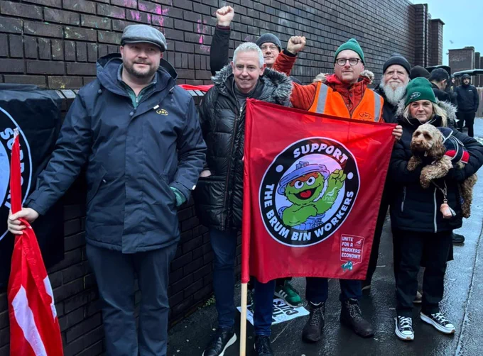 Group of RMT members standing by a brick wall holding a red flag supporting Brum bin workers with raised fists in solidarity.
