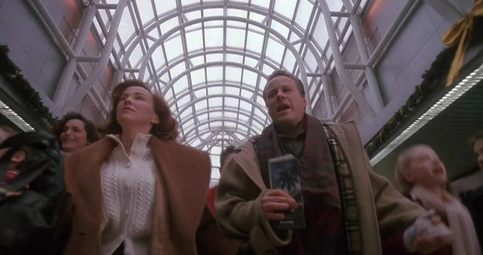 A group of people walk briskly through a busy airport terminal under a large, arched glass ceiling. Holiday decorations are visible, suggesting a festive atmosphere.