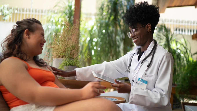 Stock photo of a woman with Down Syndrome speaking to a physician. Both are seated across from one another and smiling. The patient is wearing an orange tank top and white shorts, the physician is wearing a white coat, stethoscope, badge, and glasses and is holding a tablet. 