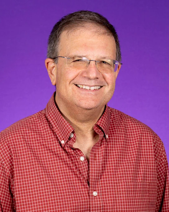 An East Carolina University faculty member wearing a red checkered shirt smiles at a camera in front of a purple photo backdrop.