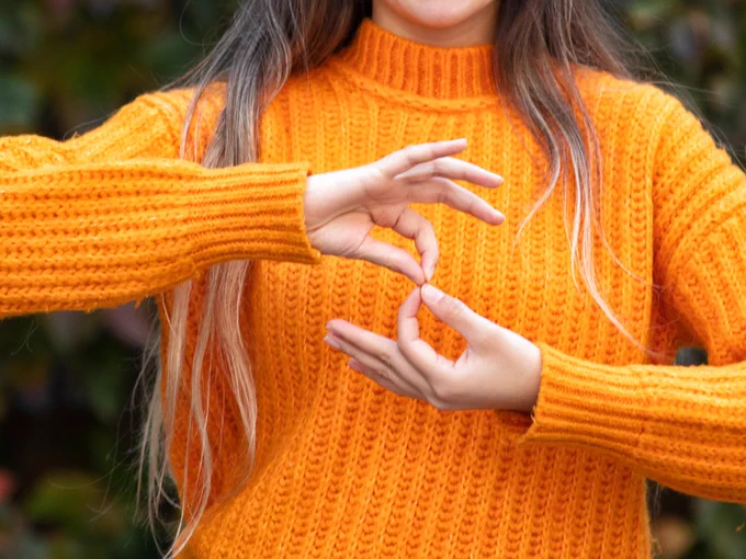 A person wearing an orange sweater uses sign language to form a symbol with their hands against a blurred background.