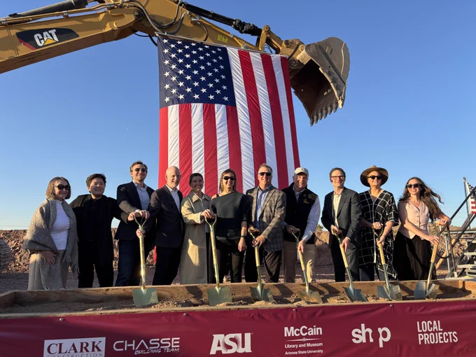 Design and construction professionals dig shovels into the ground in front of a American flag hanging from construction equipment during ceremony for the McCain Library and Museum in Tempe.