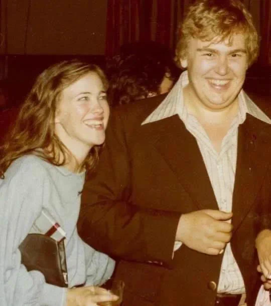 Catherine O'Hara and John Candy standing close together at what appears to be an indoor gathering. Catherine O’Hara is on the left, wearing a light blue sweater and smiling while looking to her right. She is holding a small glass in one hand and a clutch-style purse under her arm. John Candy is on the right, wearing a dark jacket over a light, open-collared shirt, smiling directly toward the camera. The background is dimly lit, with other people partially visible behind them.
