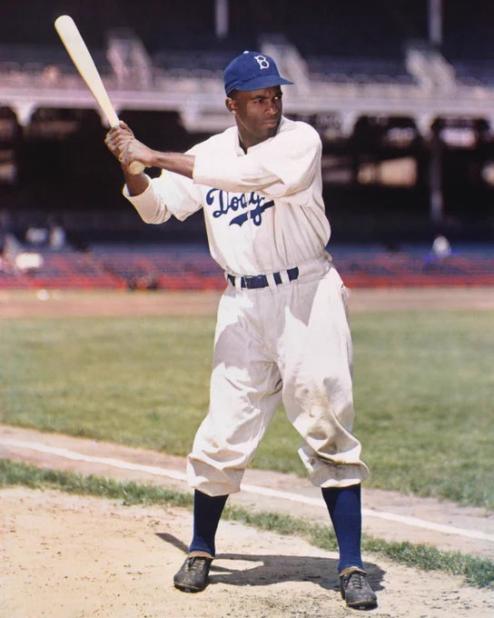A photo of Jackie Robinson standing in foul territory in a baseball stadium. He is wearing a home white Brooklyn Dodgers uniform and blue hat.