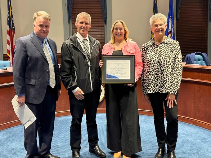 Four people stand together in a formal setting. One person holds a framed certificate. An American flag and state flags are displayed in the background, indicating a government or official venue.