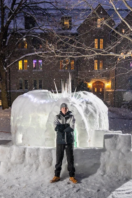 A person stands in front of a snow igloo with his arms crossed. The igloo has icicle decorations on top. Snow is falling, and a large building with lit windows is in the background.