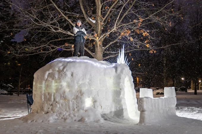 A person stands atop a large igloos set in a snowy, tree-filled space at night. Snowflakes are gently falling, illuminated by subtle ambient lighting.
