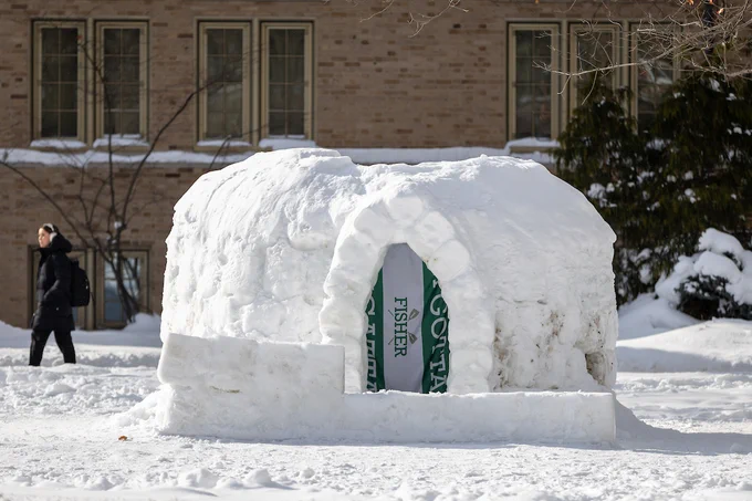 Outdoor scene featuring a snow igloo in front of a brick building with rectangular windows. The entrance of the igloo is marked by a "Fisher" banner. Snow covers the ground, and a person carrying a backpack walks past in the background.