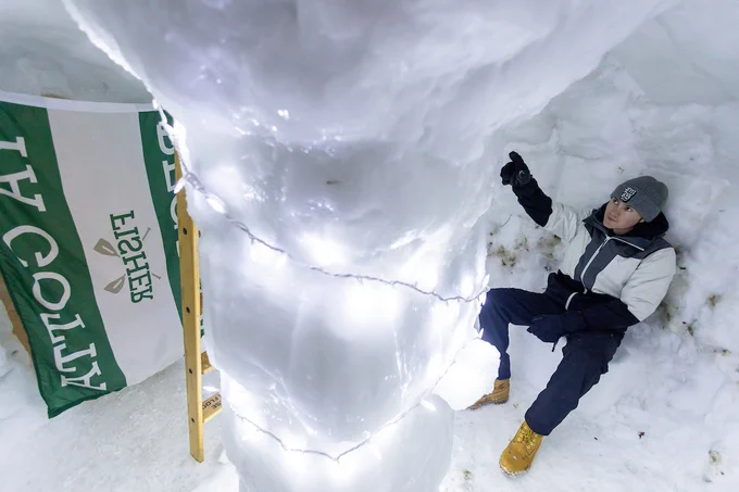 Inside an igloo, a person in a winter outfit sits next to a snow pillar wrapped with string lights. A large outdoor flag with green text is partially visible. Snow surrounds the entire scene.