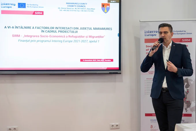 The SIRM project presentation displayed on the screen, with the speaker standing to the right of it, speaking into the microphone against the backdrop of the project roll-up.