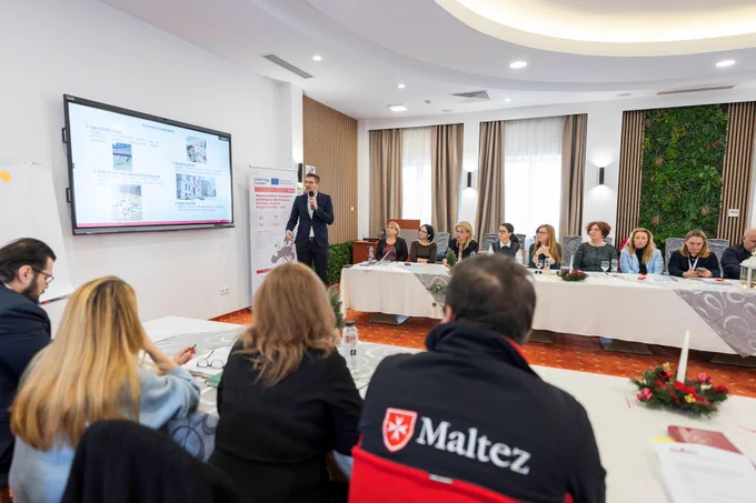 Participants sitting at tables and looking towards a screen displaying a presentation. A speaker with a microphone is standing in front of the screen, with a SIRM project roll-up visible behind them.