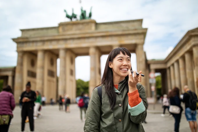 Young woman speaksing into her mobile phone, in front of the Brandenburg Gate in Berlin on a cloudy day.