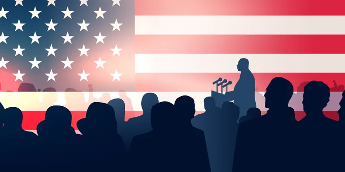 Silhouette of a person speaking at a podium, with an audience in the foreground, against a backdrop of the American flag.