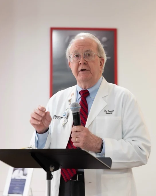 Dr. Taylor, in a white lab coat, speaking at a lectern with a microphone in a room with red framed artwork and informational posters.