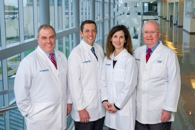 Four medical professionals in white lab coats, standing together in a brightly lit hospital corridor, with large windows showing a view outside. They all wear badges and two have stethoscopes.