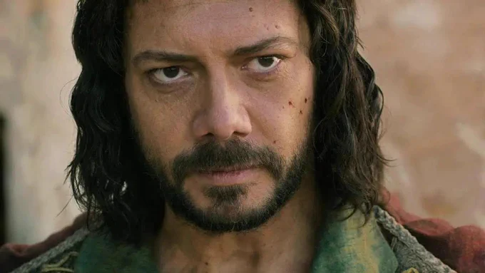 Close-up portrait of Álvaro Morte as Logain Ablar, staring directly forward with an intense, defiant expression. He has long, dark, wavy hair, a full beard, and visible dirt and small cuts on his face. He wears layered, worn clothing in muted green and red tones, with a softly blurred stone background behind him.
