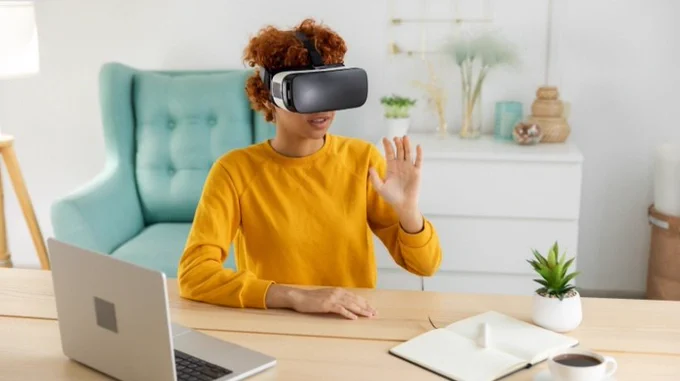Person wearing a virtual reality headset sitting at a desk with a laptop, notebook, and small plant, waving their hand in a bright room.