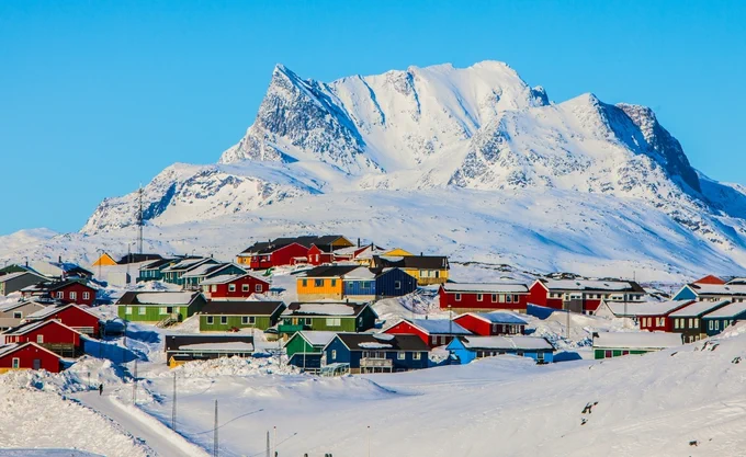 A vibrant cluster of colorful houses, painted in red, green, yellow, and blue, is nestled on a snowy landscape. The scene is set against the backdrop of a majestic, snow-covered mountain under a bright blue sky.