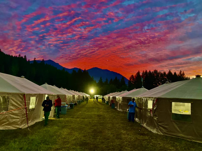 Rows of illuminated tents at the Incident Command Post line a grassy path under a vibrant red and purple sunset sky with silhouetted mountains and trees in the background. Photo by Paul Hohn. 