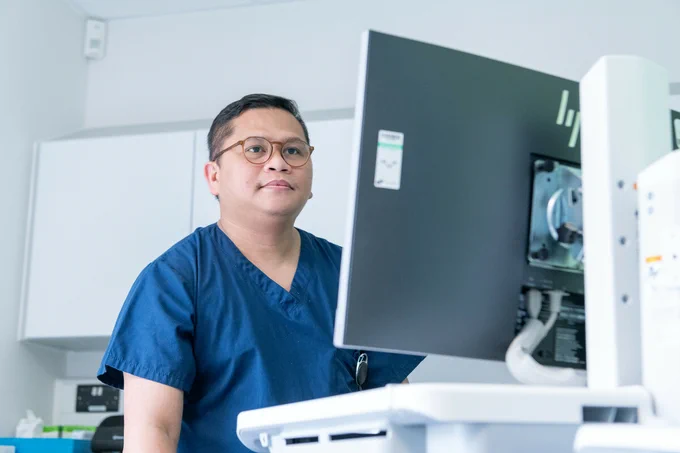 A healthcare professional in blue scrubs stands beside a computer, focused on the screen in a modern medical environment