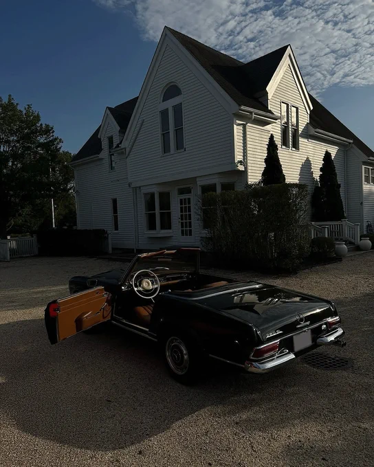 A black vintage convertible car parked in front of a large white house with a steep roof. The driver's door is open, revealing a tan interior. The house features multiple windows, shrubs, and a gravel driveway under a partly cloudy sky.